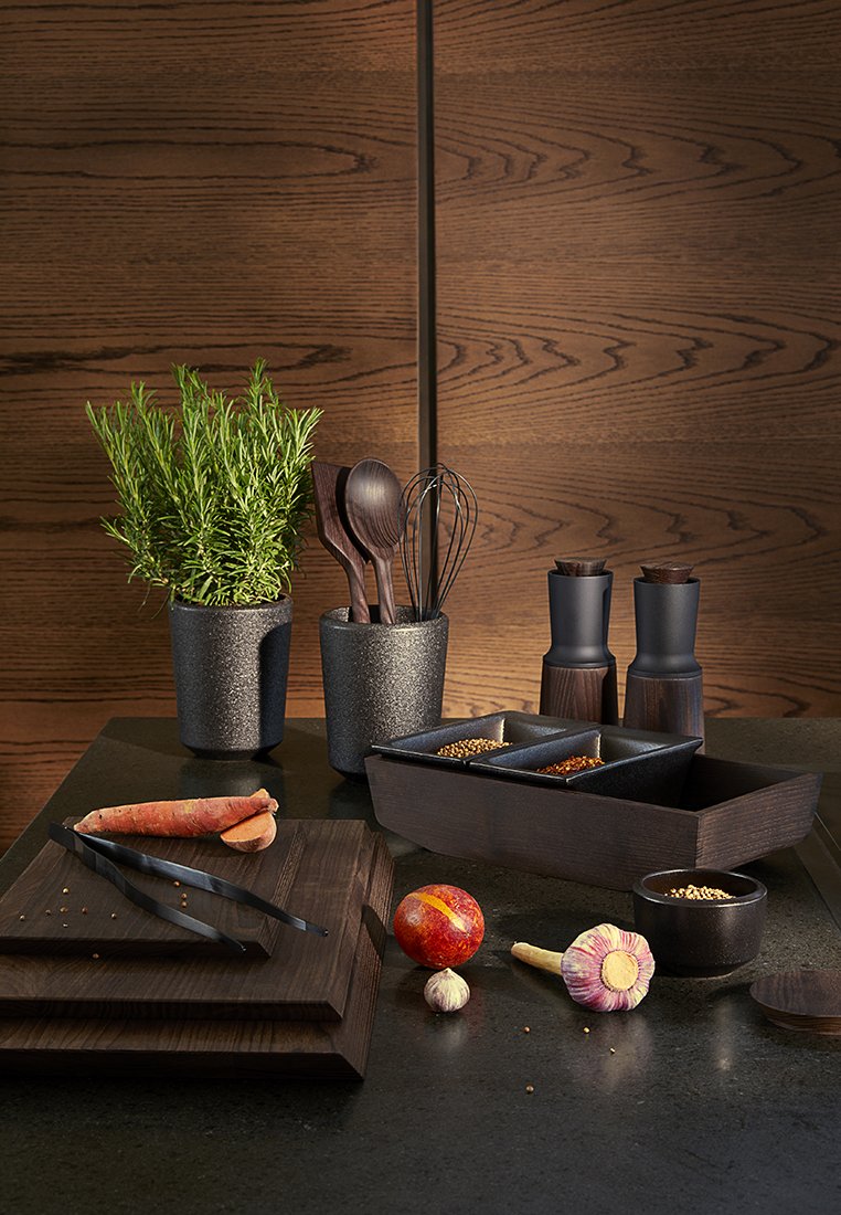 Ensemble d'ustensiles de cuisine comprenant des casseroles noires texturées, des ustensiles en bois, des contenants d'épices, des planches à découper et des herbes fraîches sur une surface sombre.