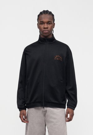 Mladý muž s pletenými vlasmi, oblečený v čiernej bunde na zips s logom „Carhartt WIP“ a svetlosivých nohaviciach na jednoduchom pozadí.