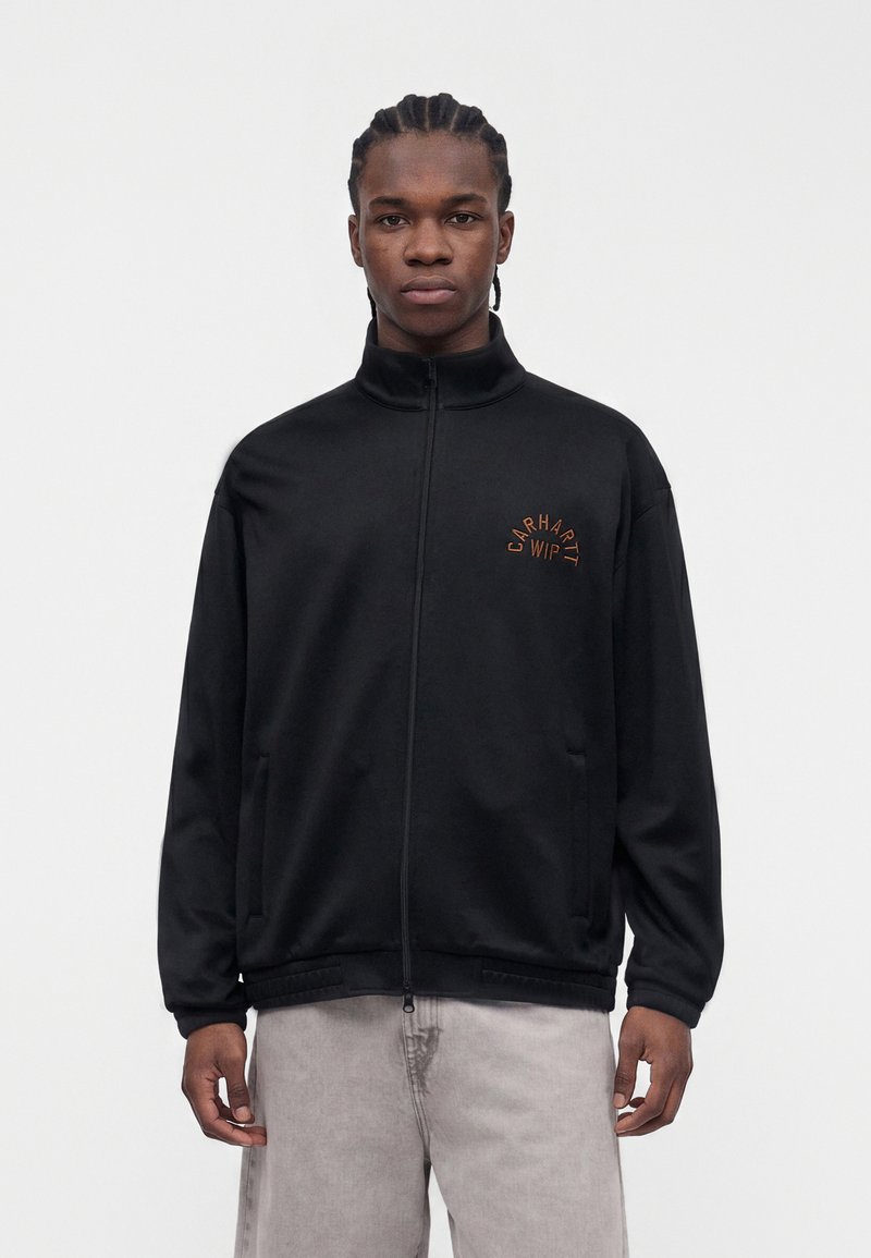 Junger Mann mit geflochtenem Haar, der eine schwarze Reißverschlussjacke mit „Carhartt WIP“-Logo und hellgraue Hosen vor einfachem Hintergrund trägt.