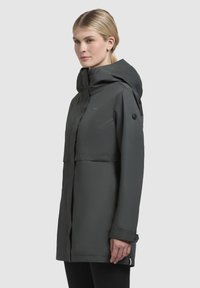 Donkergrijze waterdichte jas met een capuchon, voorzien van een voorklep en knoopdetails op de manchetten. Soepele, matte textuur met een minimalistisch ontwerp.