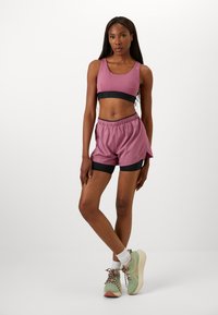 Light pink sports bra with black band, paired with light pink shorts featuring black accents. Model wears green athletic shoes with patterned laces.
