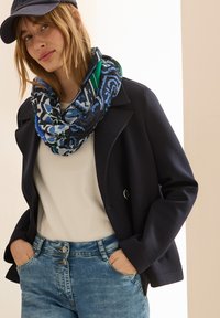 Marineblå cropped jakke med store klapper, båret over en lys bluse, suppleret med et mønstret blå-grønt infinity-scarf og lyseblå jeans.