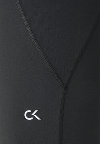 Sorte atletikleggings med elastisk materiale, glat tekstur og et hvidt CK-logo nær kanten med kontrasterende syningsdetaljer.