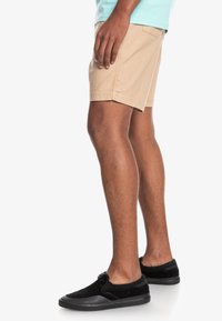 Beige bomullsshorts med rak skärning och sidfickor, visade på en person som bär svarta slip-on skor.