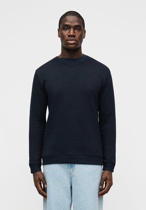 Homme portant un sweat-shirt bleu marine uni et un jean bleu clair, debout face à un fond clair uni.