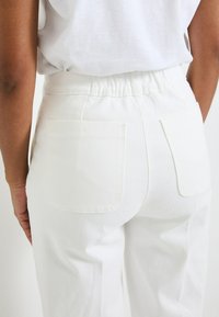 Pantalones de mezclilla blanca con cintura elástica, que presentan dos bolsillos traseros y una tela suave y texturizada. Diseño limpio y moderno.