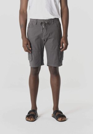 Férfi, aki szürke, zsinóros derekú haszongatya rövidnadrágot és fekete szandált visel, egy egyszínű szürke háttér előtt áll.