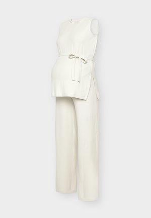 Bezrukávový krémový pletený tehotenský top s bočnými rozparkami a s kúskom na zaväzovanie v páse v páre so širokými pletenými nohavicami v rovnakom dizajne.