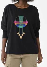 Chemise en coton noire à manches longues avec un motif géométrique brodé multicolore, orné de franges et d'accents circulaires sur la poitrine.