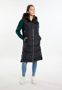 Schwarze Steppweste mit Kapuze, mit einem Reißverschluss vorne, Seitentaschen und einem gesteppten Muster. Kombiniert mit einem grünen strukturierten Pullover und Jeans.