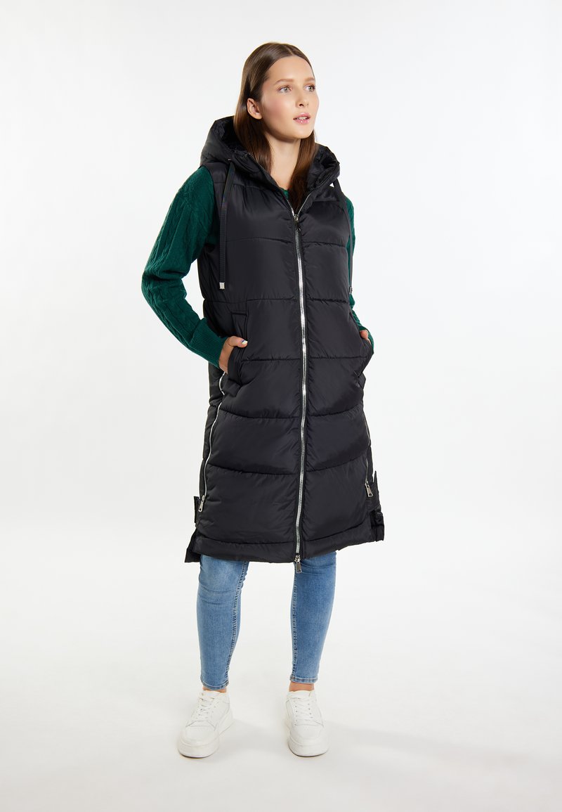 Schwarze Steppweste mit Kapuze, mit einem Reißverschluss vorne, Seitentaschen und einem gesteppten Muster. Kombiniert mit einem grünen strukturierten Pullover und Jeans.
