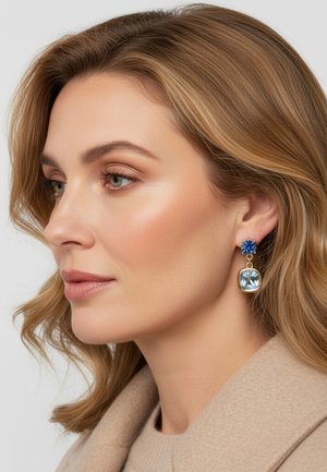 Vrouw met lichtbruin haar die blauwe oorringen met edelstenen en een beige jas draagt, gericht naar links tegen een effen achtergrond.