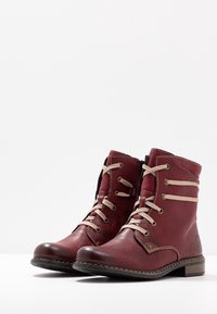 Røde læder ankelstøvler med beige snørebånd, rund tå og en lille hæl. Har sidesøm og et logoaccent på den ydre side.