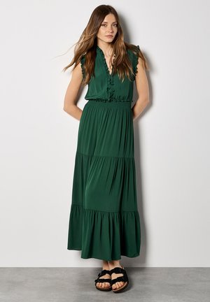 Mujer de pie con las manos detrás de la espalda, vistiendo un vestido largo sin mangas verde oscuro con volantes y sandalias negras con hebilla, frente a una pared blanca.