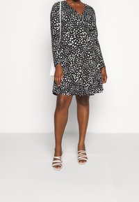 Czarna sukienka w białe cętki, z długimi rękawami i dekoltem w serek. W połączeniu z białymi sandałami na cienkich paskach. Prosty krój i rozkloszowany fason.