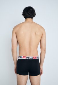 Boxer-briefs noirs avec une large ceinture blanche portant le logo rouge "DIESEL", portés par un mannequin aux cheveux bouclés, montrant la vue de dos.