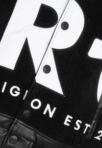 Czarna i biała kurtka z miękkim, teksturowanym materiałem typu fleece, skórzanymi akcentami, dużym logo oraz czarnymi zatrzaskami.