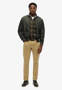 Groene bomberjack over een geruite shirt, khaki broek en zwarte schoenen. Casual stijl met zakken op de jas en riemaccenten.