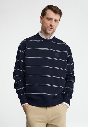 Uomo con capelli castani corti che indossa una felpa a righe blu navy sopra una camicia bianca, pantaloni beige, in piedi con le mani in tasca.