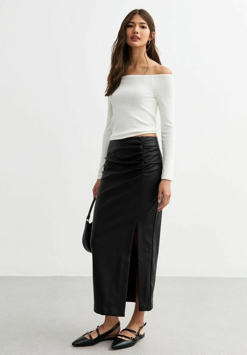 Vit off-shoulder topp med långa ärmar, kombinerad med en svart lädermaxikjol med rynkning och en sidöppning. Svarta platta skor fulländar looken.