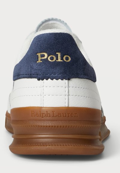 Zapatilla blanca con un talón de ante en color azul marino. Presenta el logo dorado "Polo" y "Ralph Lauren" grabados en la suela de goma en tono beige. Detalles de costura visibles.