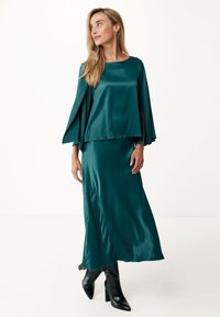 Turkusowa satynowa sukienka z opadającymi rękawami w formie peleryny i prostym krojem, zestawiona z czarnymi, wysokimi, spiczastymi kozakiem. Gładka tekstura, elegancki design.