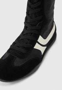 Černá sportovní bota z kombinace semiše a síťoviny, s texturovaným povrchem, plochými tkaničkami a bílým proužkem jako akcentem.