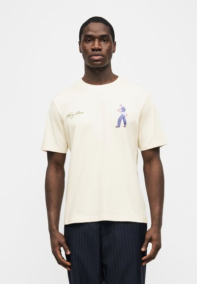 Άνδρας που φοράει κρεμ μπλουζάκι με μικρό γραφικό ενός περπατημένου ατόμου και κειμενογραφία με γράμματα στην περιοχή του στήθους, συνδυασμένο με σκούρο παντελόνι με ρίγες.