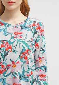Blusa de manga larga floral con una base gris y vibrantes estampados botánicos en rojo, rosa y verde. Cuello redondo, textura suave, corte relajado.