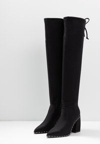 Schwarze Overknee-Stiefel aus glattem Stoff mit spitzem Schuhkopf, Blockabsatz und Nietenakzenten entlang des Absatzes und der vorderen Naht.