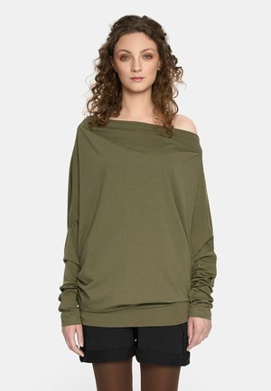 Oliwkowa bluzka na ramiączkach z długimi rękawami i marszczeniami. Połączona z czarnymi szortami i brązowymi rajstopami. Gładka tekstura materiału.