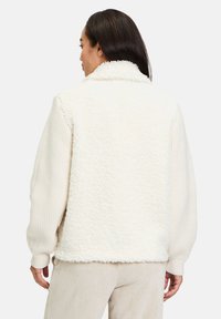 Cremefarvet fleecejakke med en tekstureret, langhåret ryg og ribstrikkede ærmer. Åben krave og ingen synlige lukninger. Blød og hyggelig fremtoning.