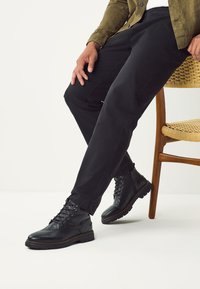 Čierne kožené členkové topánky s textúrovaným povrchom, šnúrkami a robustnými podrážkami, skombinované s tmavými nohavicami a zelenou košeľou, sediace na stoličke.