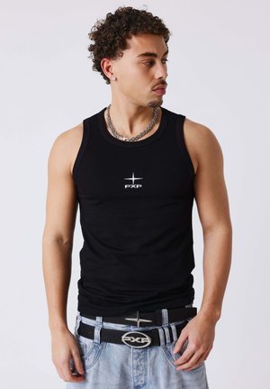 Joven con cabello rizado que lleva una camiseta negra sin mangas con un logo de estrella, collar de cadenas plateadas en capas, jeans azul claro y doble cinturón.