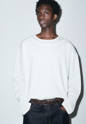 Giovane uomo con le mani in tasca, indossa una camicia bianca a maniche lunghe e larga, jeans scuri e una cintura marrone, in piedi contro uno sfondo neutro.