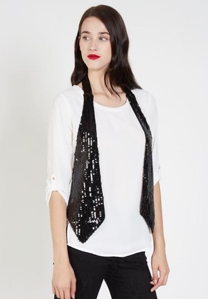 Mujer con cabello largo y oscuro, que lleva lápiz labial rojo, una camisa blanca de manga larga, pantalones negros y una bufanda negra con lentejuelas colgada alrededor de su cuello.