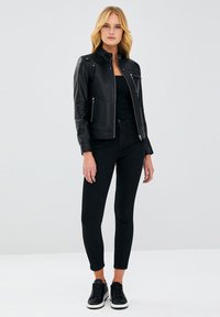 Zwarte leren motorjack met een rits aan de voorkant, een mandarin kraag en zijzakken, in combinatie met strakke zwarte broek en zwarte sneakers.