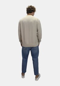Sudadera beige con puños y dobladillo acanalados, combinada con vaqueros azules. Bolsillos traseros abotonados visibles. Calzado casual completa el look.