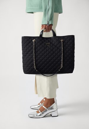 Personne tenant un sac cabas matelassé noir Guess avec des anses en chaîne, portant des chaussures argentées à talons blocs, un pantalon blanc et une veste verte avec des fermetures éclair.