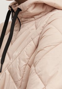 Giacca trapuntata beige con colletto alto e lacci neri, che mostra dettagli di cucitura e una texture di tessuto liscia.