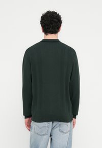 Σκούρο πράσινο πλεκτό πουλόβερ με ριβέρ λεπτομέρειες στην πλάτη, άνετη εφαρμογή και επίπεδη λαιμόκοψη. Συνδυασμένο με ανοιχτό μπλε τζιν παντελόνι.