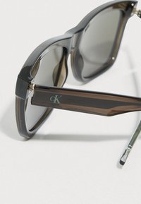 Okulary przeciwsłoneczne z ciemnoszarymi, dużymi oprawkami i gradientowymi soczewkami. Zauszniki są przezroczisto-brązowe z subtelnym logo "ck" blisko zawiasu.