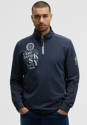 Navyblauer Langarm-Pullover mit einem halben Reißverschluss am Kragen. Verfügt über weiße Grafiken und Text auf der Vorderseite und dem linken Ärmel. Weicher Stoff mit glatter Textur.