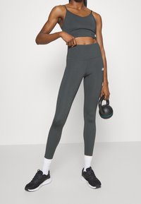 Reggiseno sportivo grigio e leggings a vita alta abbinati, design aderente, texture liscia, con un logo sulla gamba; persona che tiene un kettlebell.