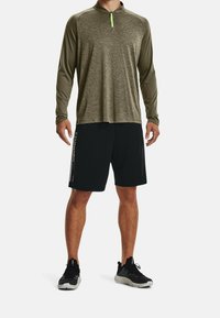 Langärmeliges, olivgrünes Sportshirt mit Reißverschlusskragen, kombiniert mit schwarzen Shorts mit Seitenstreifen. Schwarze Sneakers vervollständigen das Outfit.