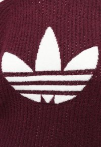 Burgundi színű kötött anyag, amelyen egy nagy fehér Adidas logo található. A dizájn három csíkot és levél formájú motívumot tartalmaz, texturált felülettel.