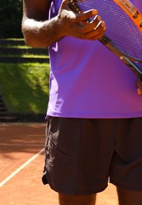 Lila träningsskjorta och bruna shorts, håller en tennisracket, visar strukturert tyg, korta ärmar och en avslappnad design.
