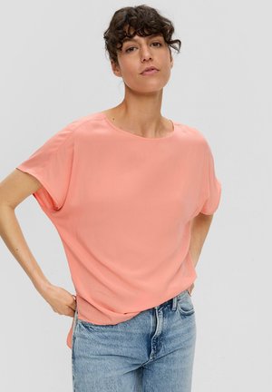 Korte mouwen, losvallend T-shirt van licht koraalkleurige stof. Ronde halslijn, zachte textuur, subtiele val, gecombineerd met lichtblauwe denim jeans.