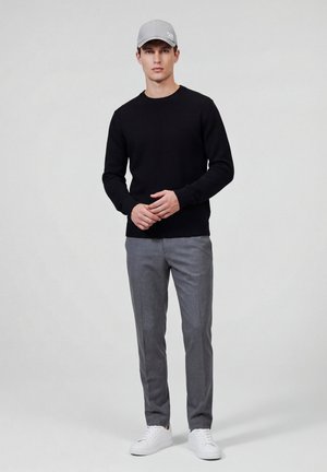 Joven de pie, vestido con un suéter negro, pantalones grises, zapatillas blancas y una gorra gris clara, contra un fondo liso y claro.