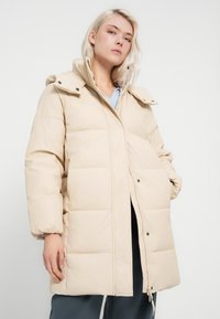 Jovem mulher vestindo um casaco acolchoado bege claro com gola alta, posando contra um fundo branco simples.